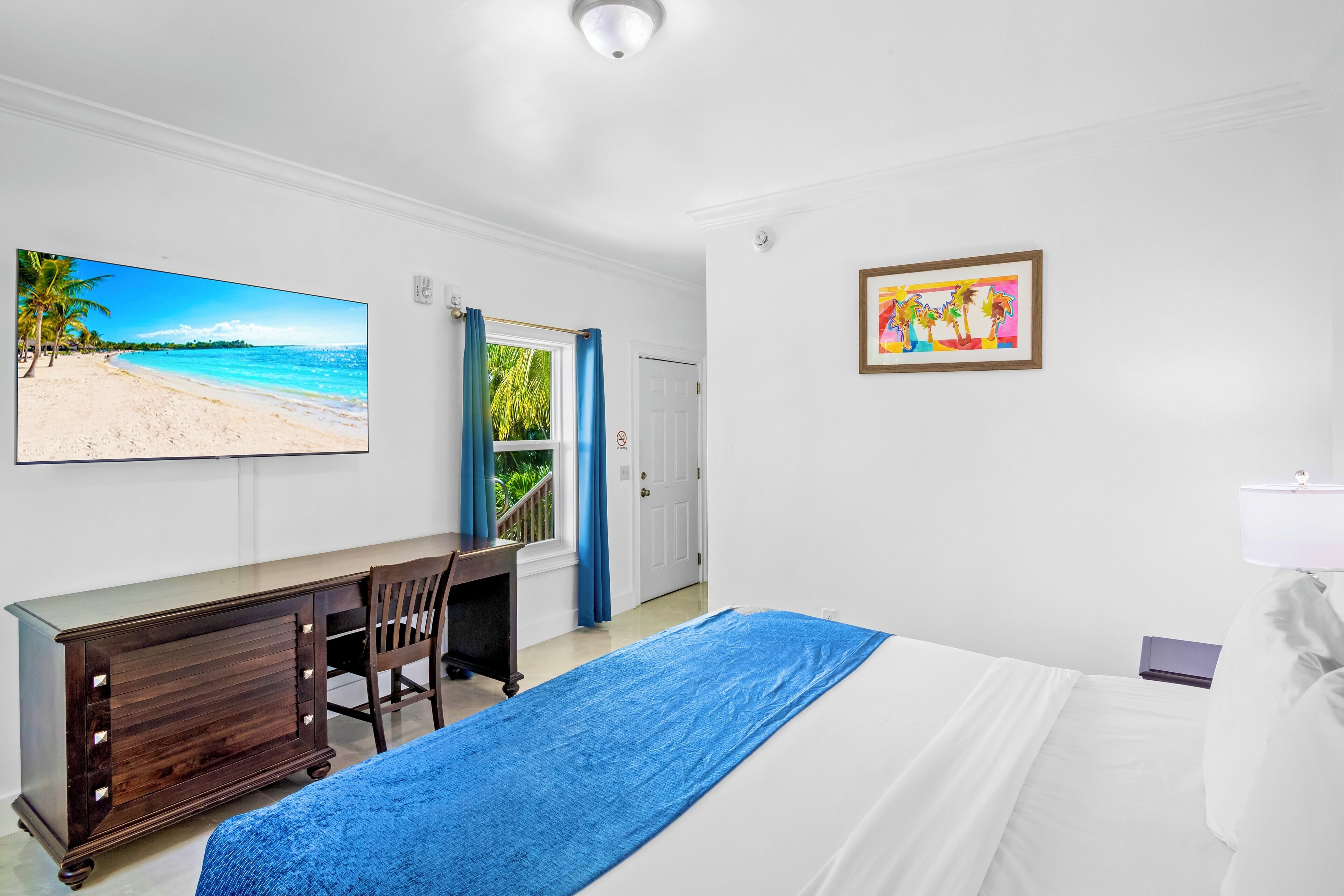 A hotel room with a bed, a desk, a chair, a TV displaying a beach scene, a framed picture, and a window with curtains.