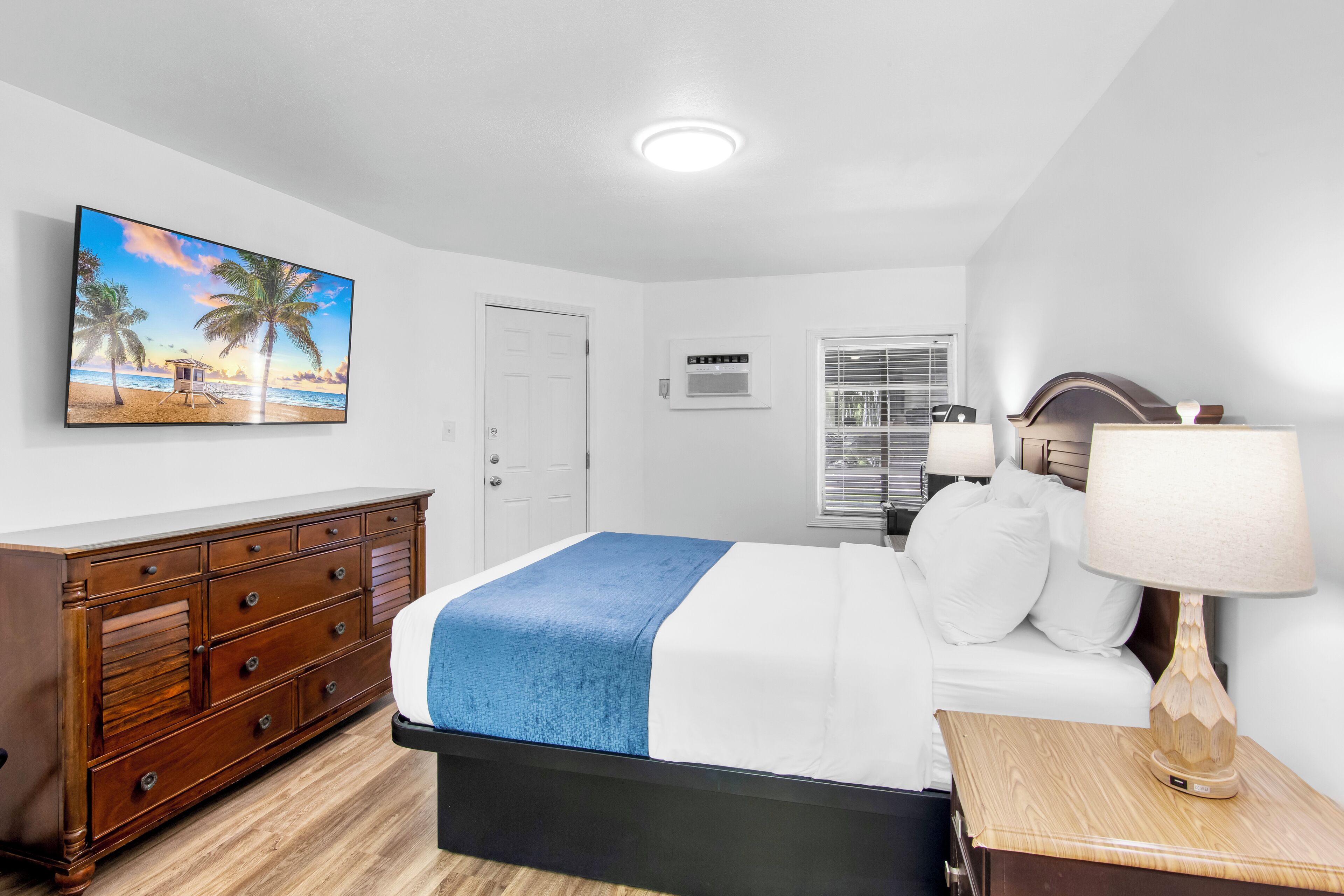 A bedroom with a bed, a wooden dresser, a painting of a beach scene, a window with blinds, and a lamp on a bedside table.