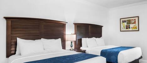 A hotel room with two beds, wooden headboards, white bedding, a ceiling fan, a nightstand with a lamp, and a framed picture on the wall.