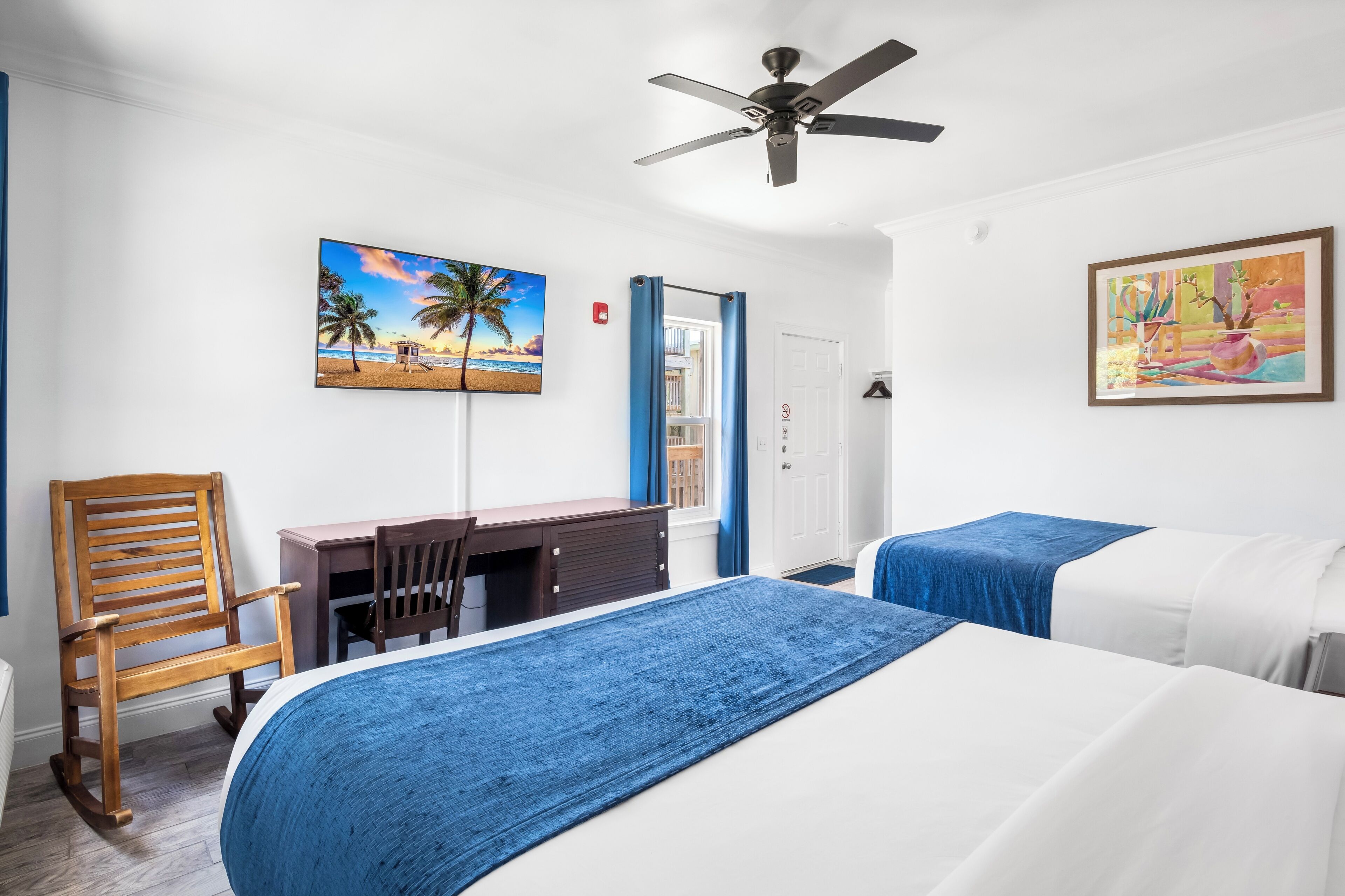 A hotel room with two beds, a desk, a chair, and a ceiling fan.