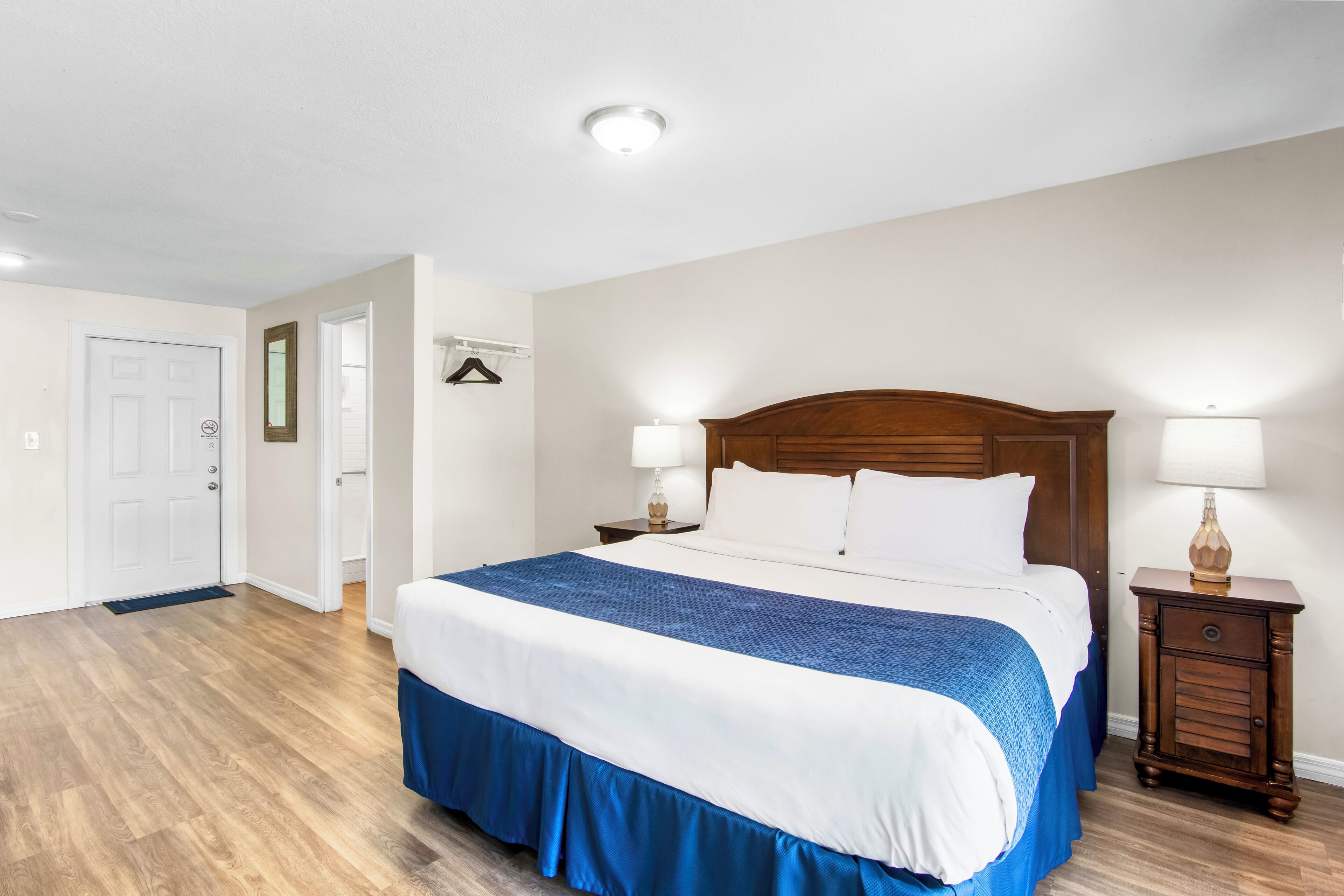 Quarto de hotel com uma cama grande, cabeceira de madeira, duas mesas de cabeceira com abajures, uma porta e um corredor.