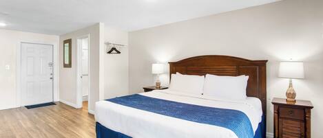 Quarto de hotel com uma cama grande, cabeceira de madeira, duas mesas de cabeceira com abajures, uma porta e um corredor.