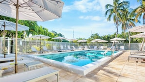 Un espace piscine agrémenté de chaises longues et de parasols, entouré de palmiers et d’une clôture blanche.