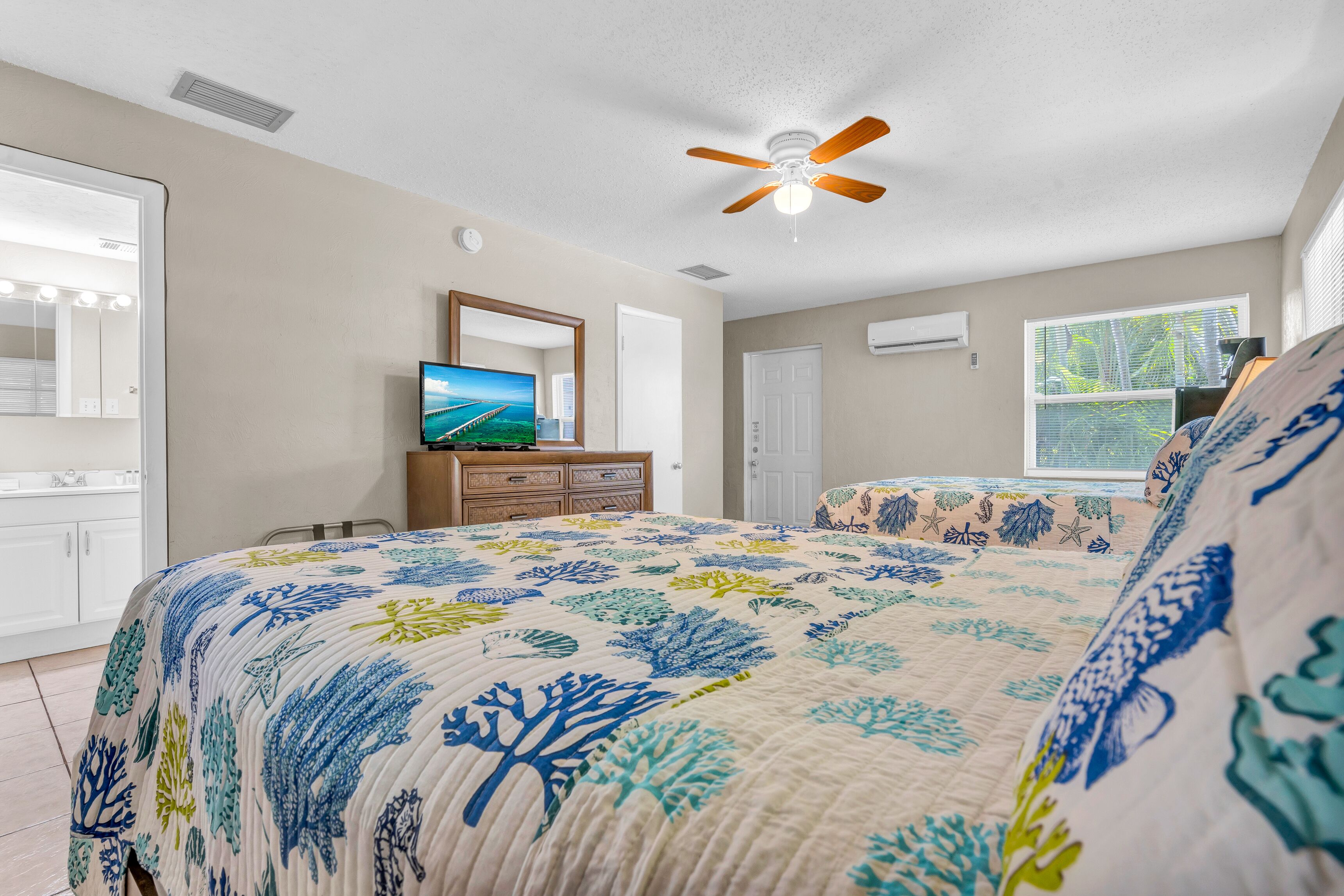 A bedroom with a bed, a ceiling fan, a TV, a dresser, a window, and a bathroom with a mirror and lights.