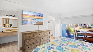 Une chambre à coucher avec un lit, une commode, une grande fenêtre et un tableau représentant un coucher de soleil sur l’océan.