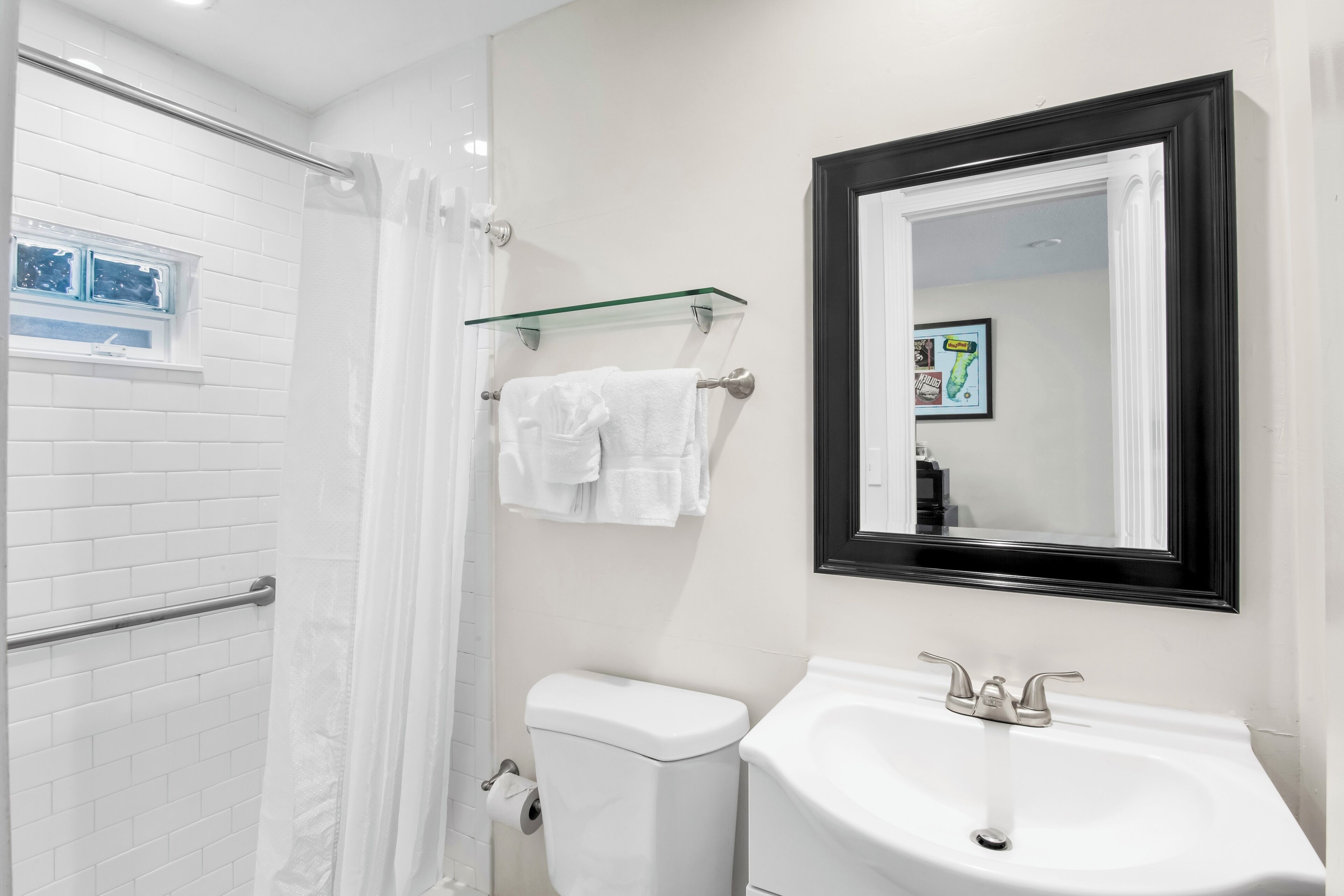 Un baño con inodoro blanco, repisa de vidrio con toallas, lavabo con espejo y cortina de ducha.