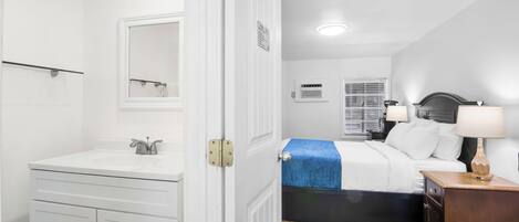 Un dormitorio con cama, mesita de noche, lámpara y un baño con lavamanos y espejo.