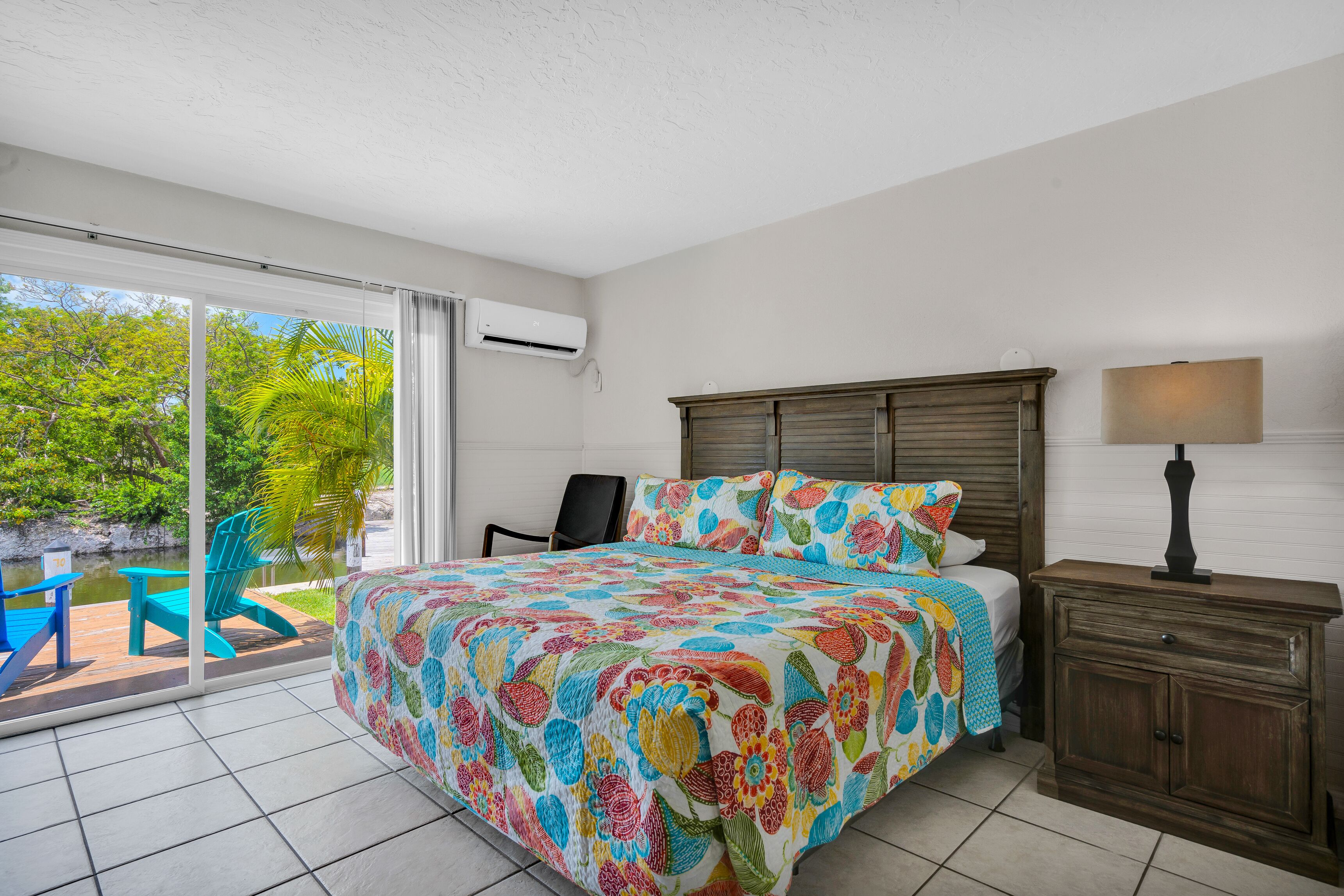 Un dormitorio con una cama grande, una mesita de noche, una silla y vistas a una piscina y vegetación al exterior.