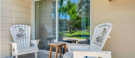 Two white Adirondack chairs with a bird design, a small wooden stool, and a white table on a balcony with a glass door.
