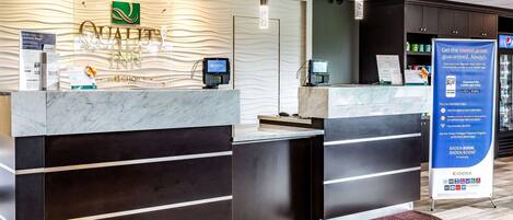 Zona de recepción de un hotel con diseño moderno, que incluye un mostrador de recepción de mármol, un cartel de Choice Hotels y un expositor promocional del programa Badda Book.