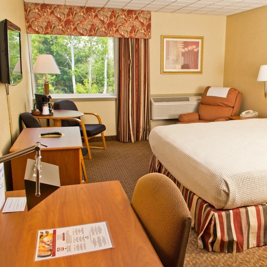A hotel room with a bed, a desk, a chair, a lamp, a telephone, a window with curtains, a picture on the wall, and a menu on the desk.