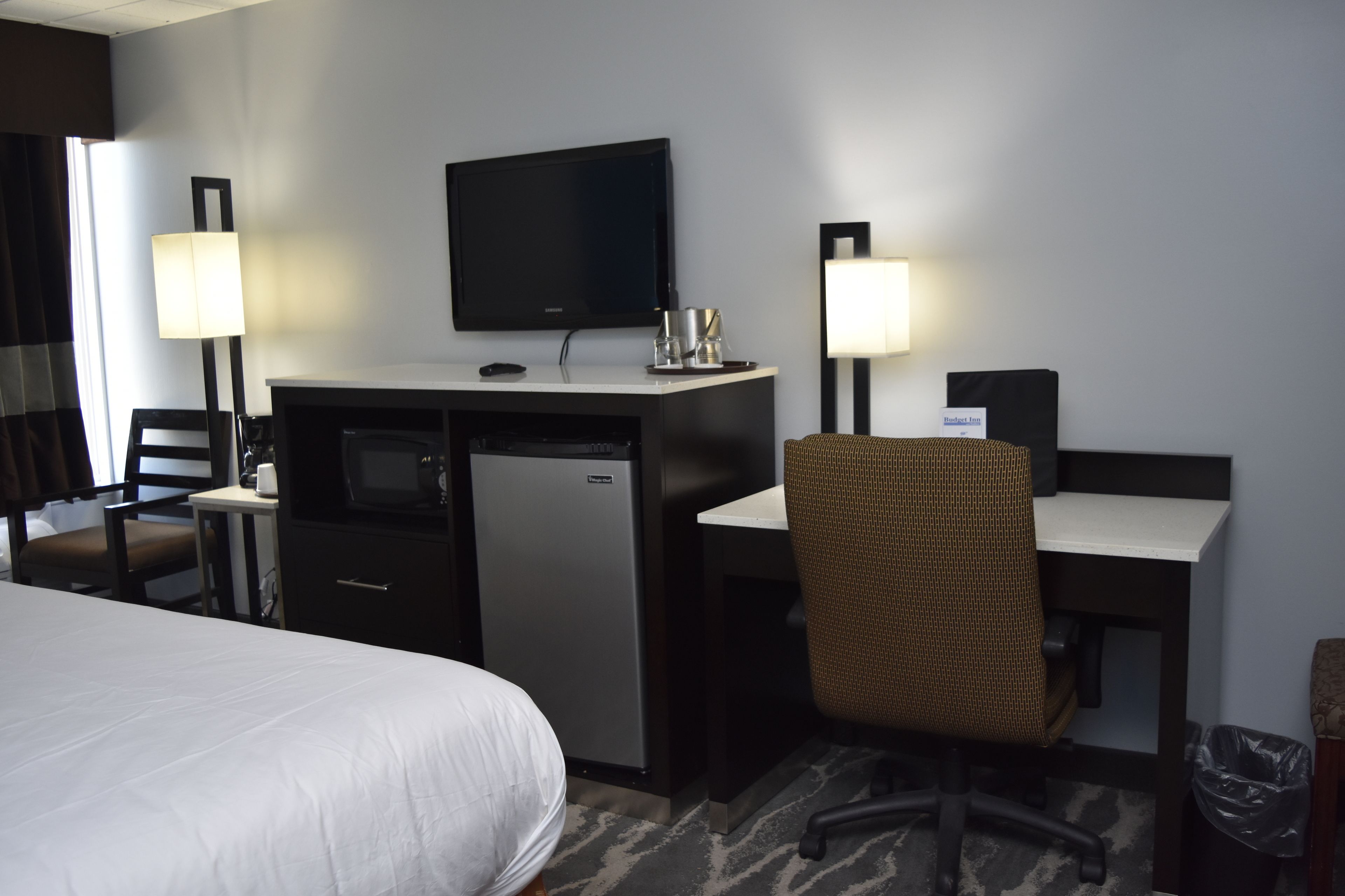 Ett hotellrum med en säng, ett skrivbord, en tv, ett kylskåp, en stol och en papperskorg.