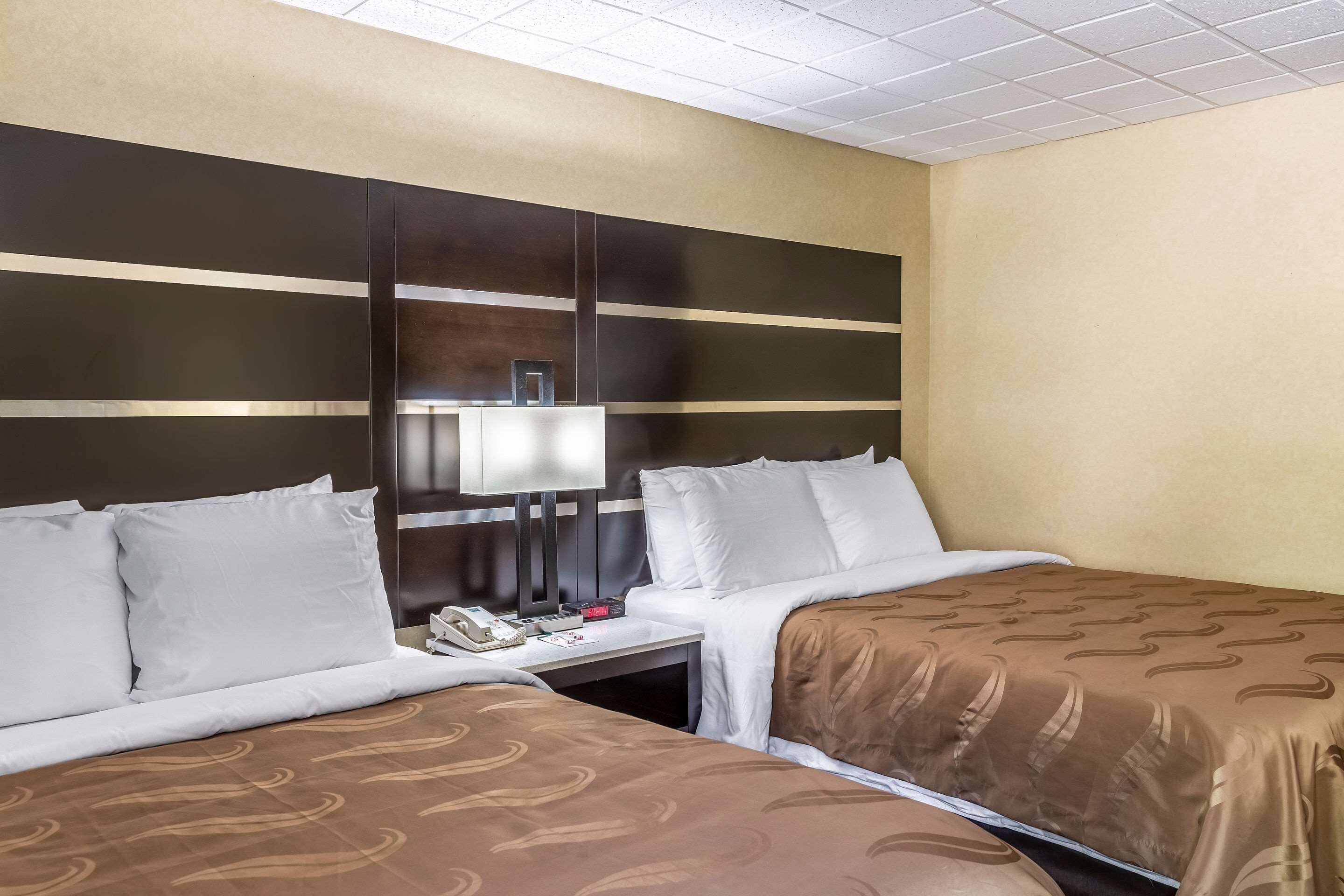 A hotel room with two beds, a nightstand with a phone and a lamp, and a large wall panel.