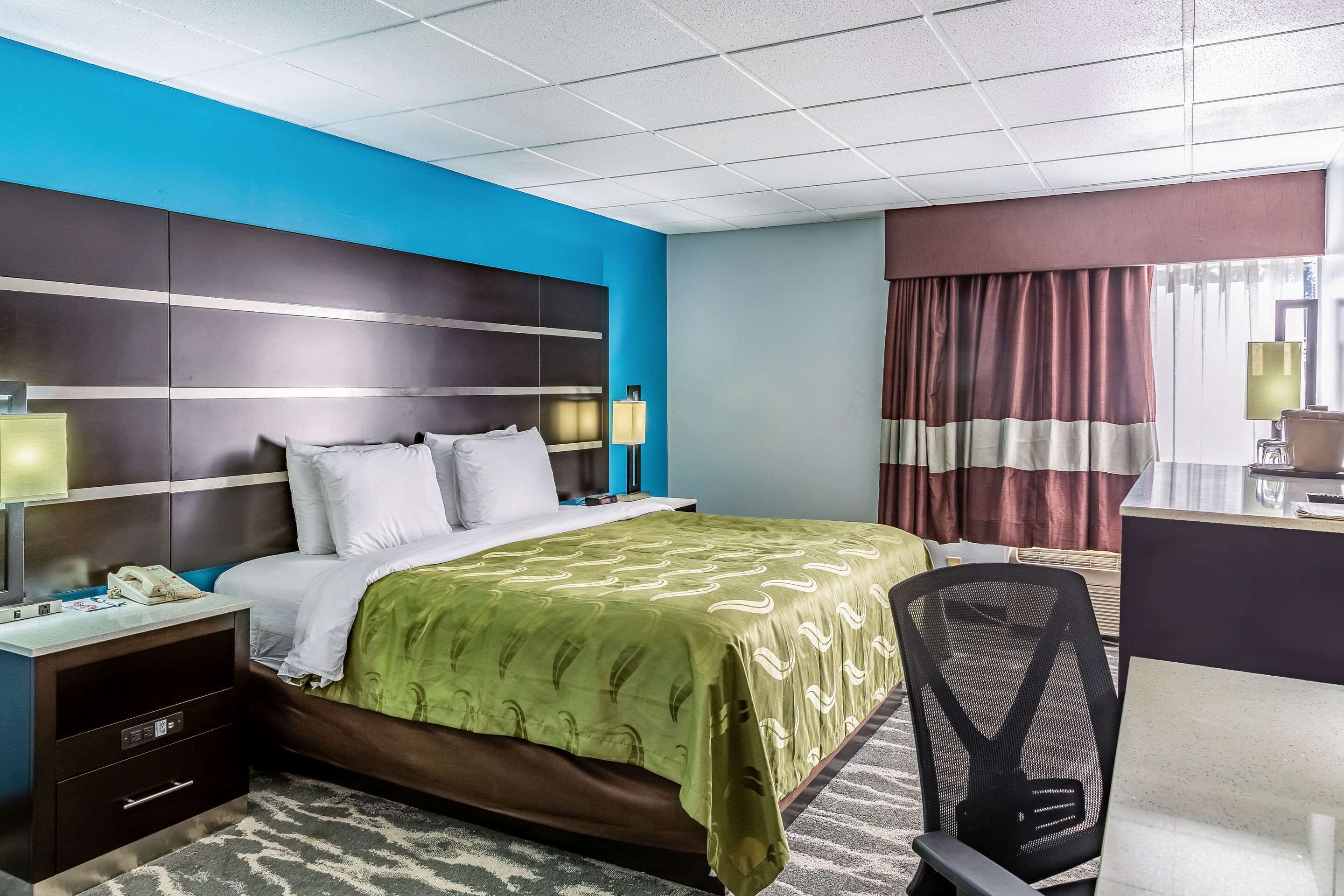 A hotel room with a bed, desk, chair, and a large window with curtains.