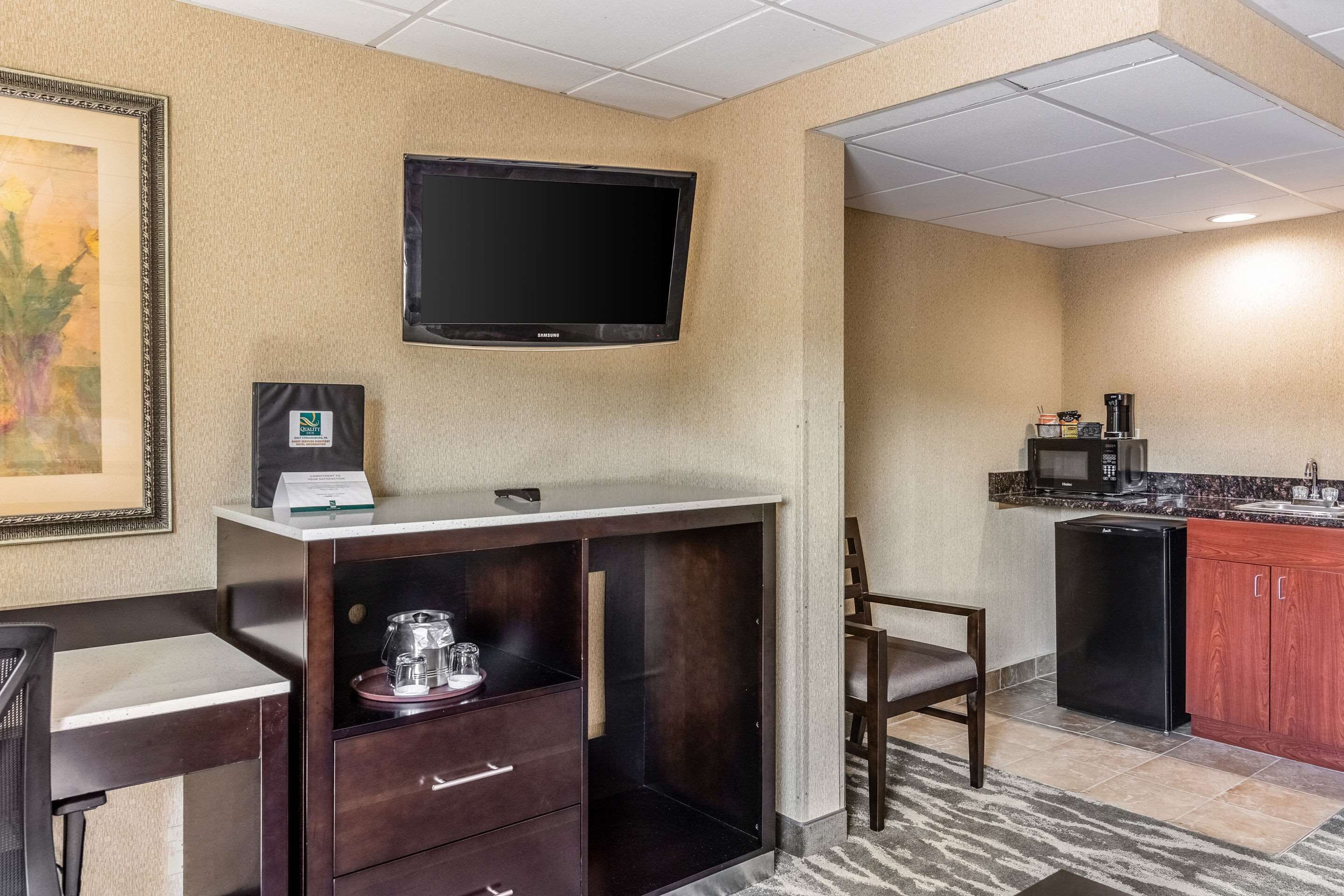 A hotel room with a kitchenette, a flat-screen TV mounted on the wall, a framed certificate on the desk, and a chair.