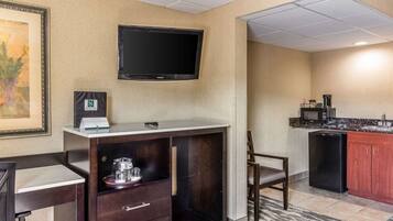 Quarto de hotel com cozinha compacta, TV de tela plana na parede, um certificado emoldurado na mesa e uma cadeira.
