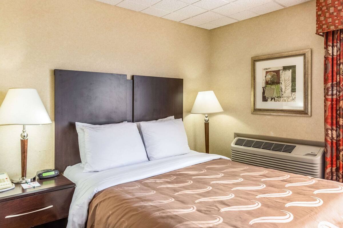 A hotel room with a bed, two bedside lamps, a nightstand with a phone and a notepad, a framed picture on the wall, and a wall-mounted air conditioner.