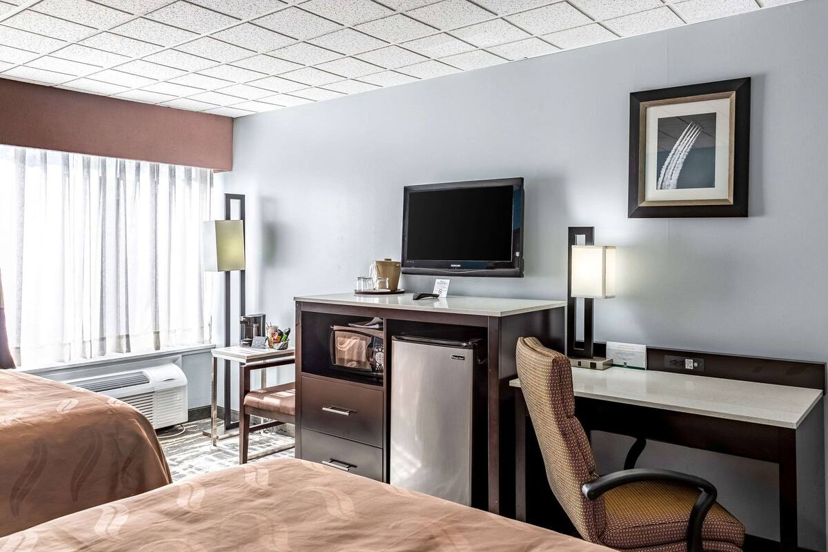 A hotel room with a bed, a desk with a computer, a refrigerator, a television, and a window with curtains.