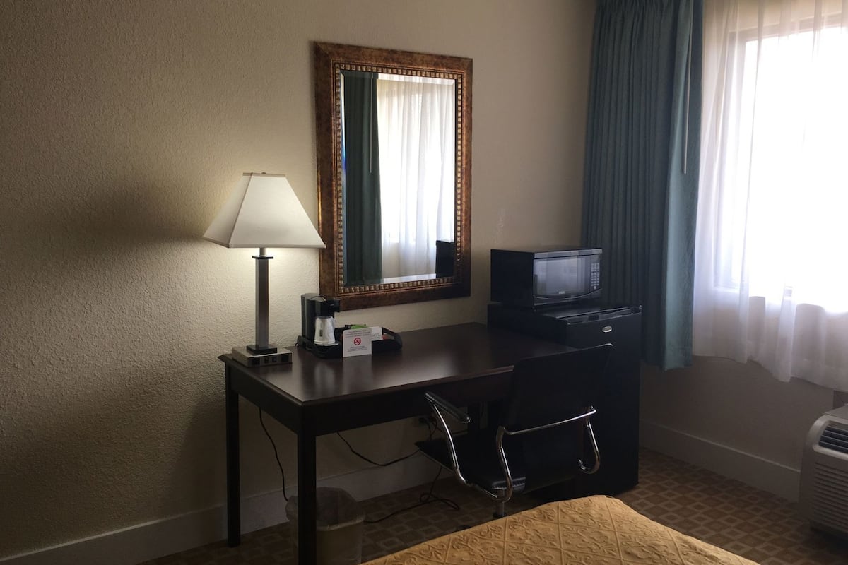 A hotel room with a desk, chair, lamp, mirror, microwave, and air conditioning unit.