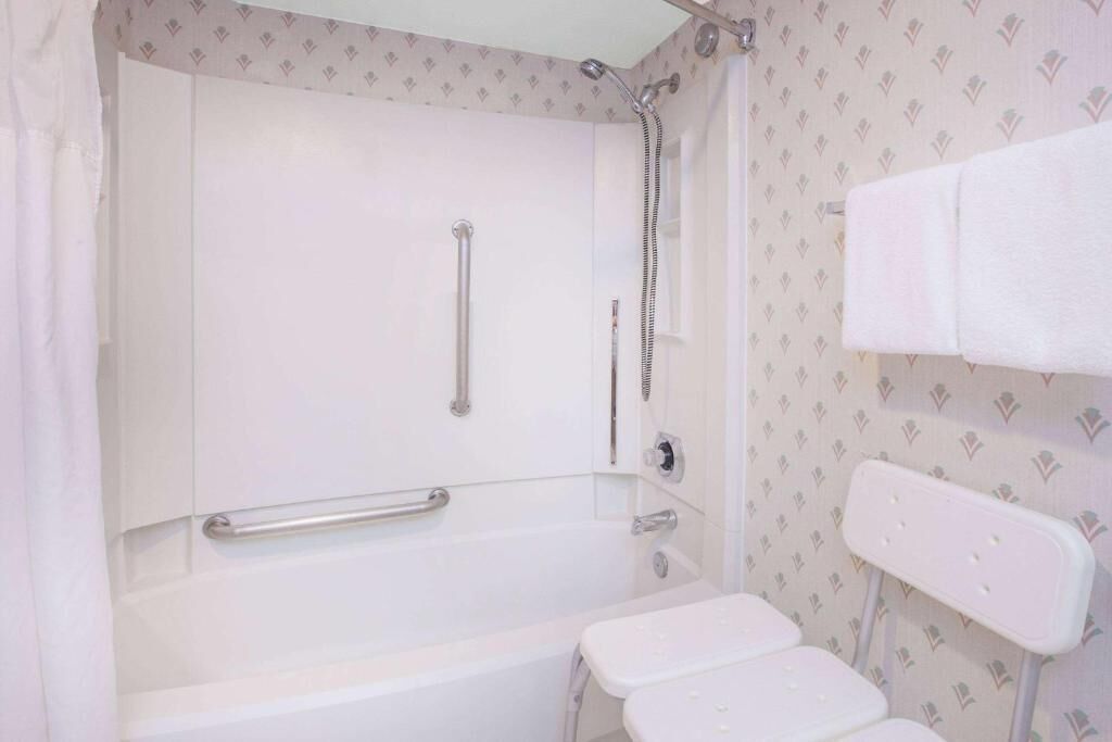 A bathroom with a bathtub, a shower, a handrail, and a white chair.
