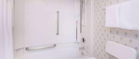 A bathroom with a bathtub, a shower, a handrail, and a white chair.