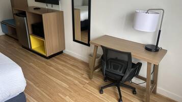 Habitación de hotel con cama, escritorio y silla, televisor, espejo, cesto de basura y lámpara.