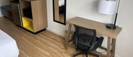 Habitación de hotel con cama, escritorio y silla, televisor, espejo, cesto de basura y lámpara.