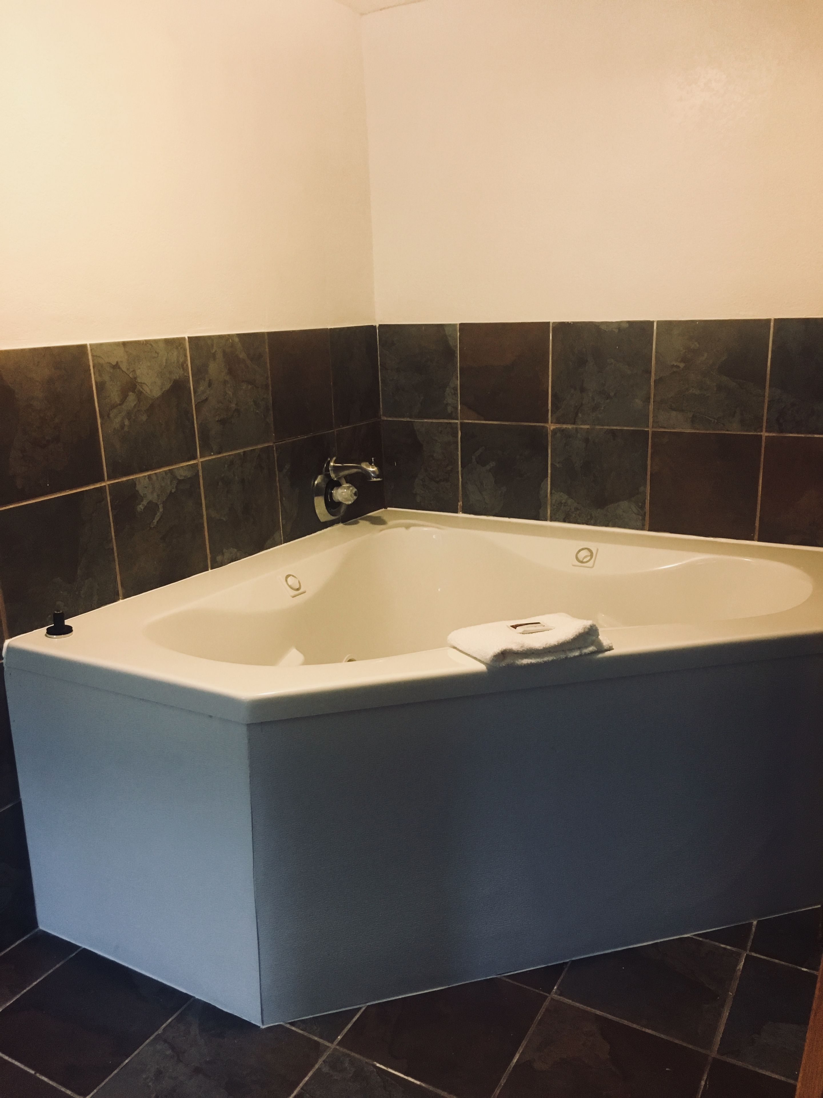 Una bañera de hidromasaje en esquina con interior blanco, paredes de azulejos negros y piso oscuro.