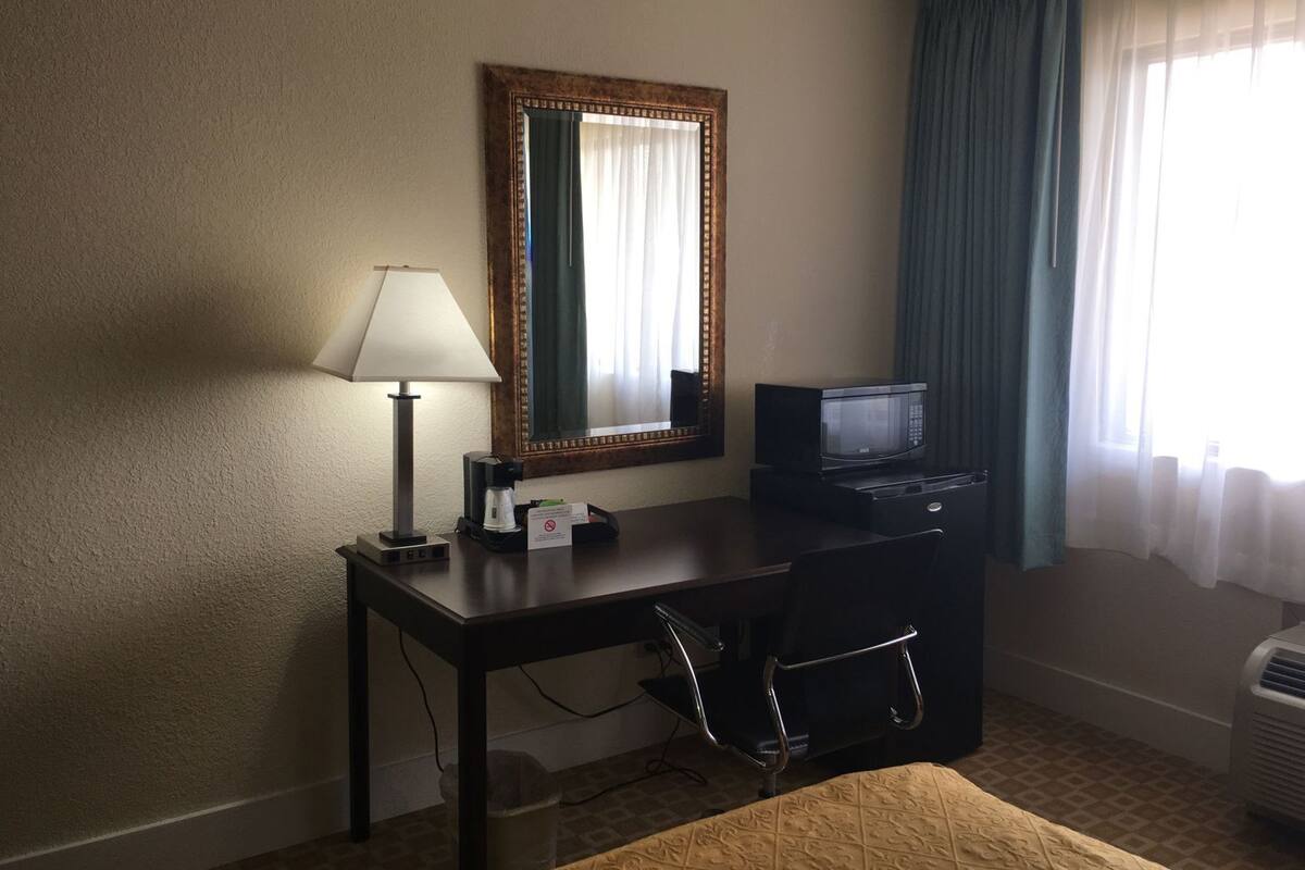 A hotel room with a desk, chair, lamp, mirror, microwave, and air conditioning unit.