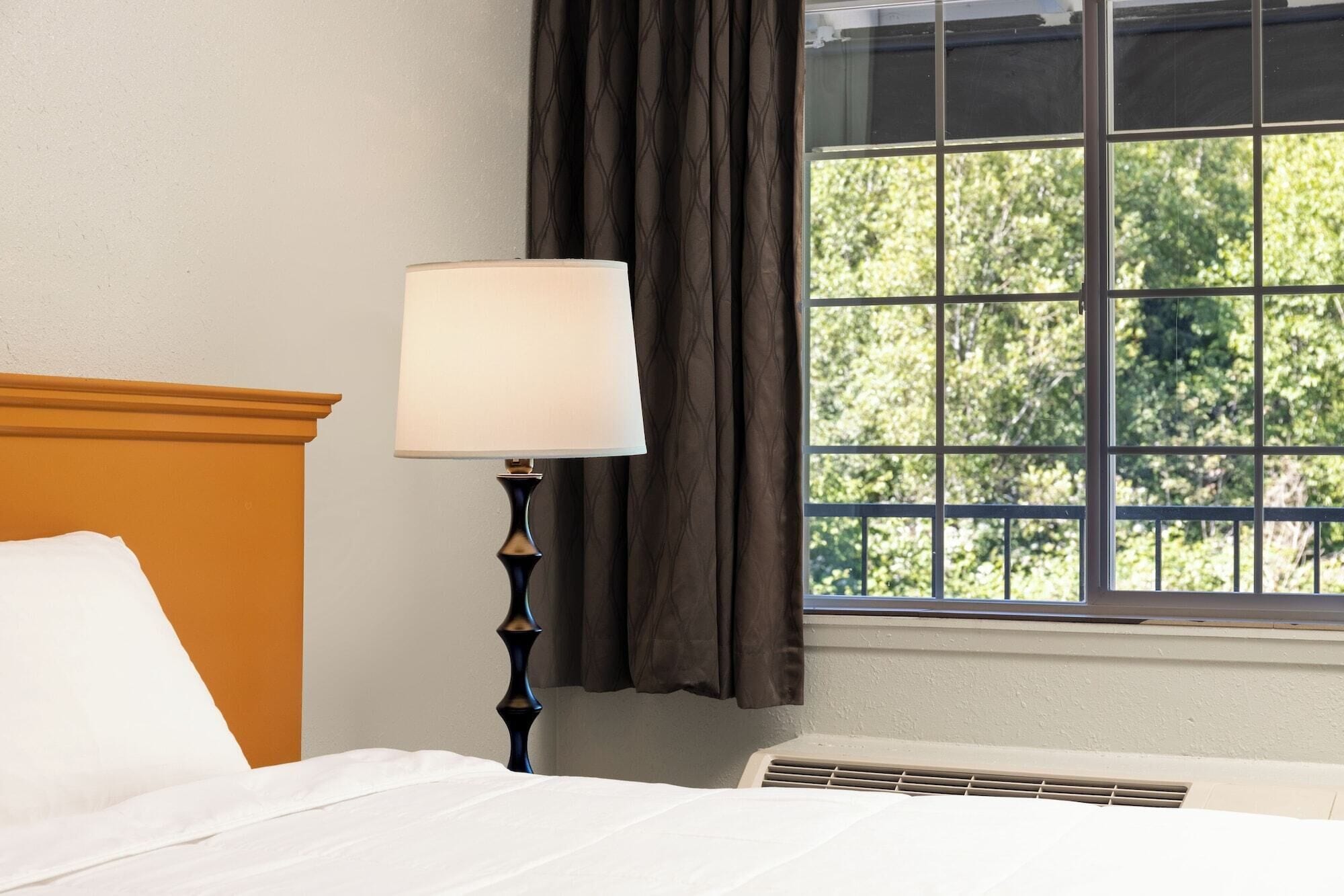 A hotel room with a bed, a nightstand with a lamp, a window with curtains, and an air conditioning unit.