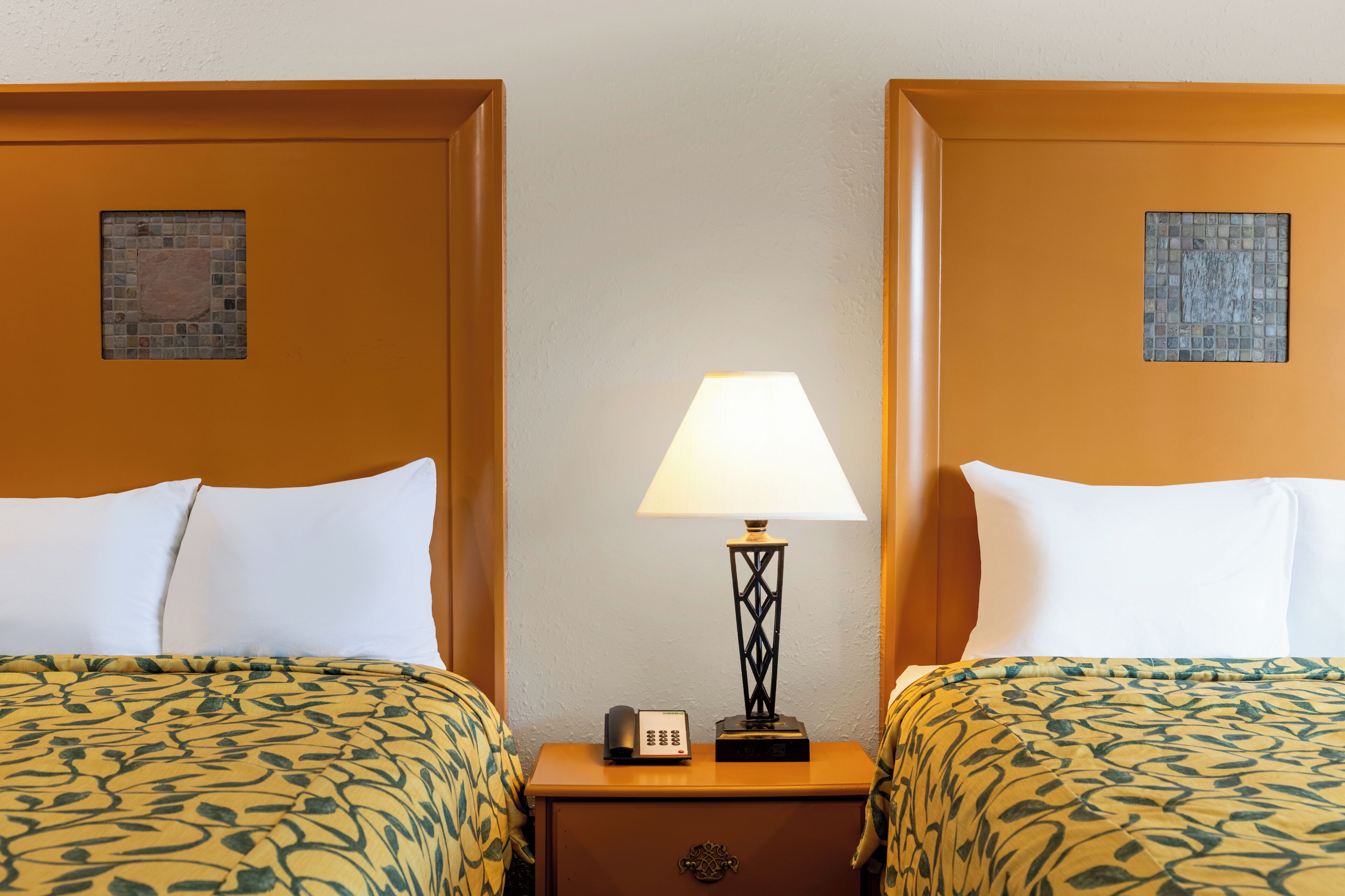 A hotel room with two beds, each with a patterned bedspread, a bedside table with a lamp, and a small clock.