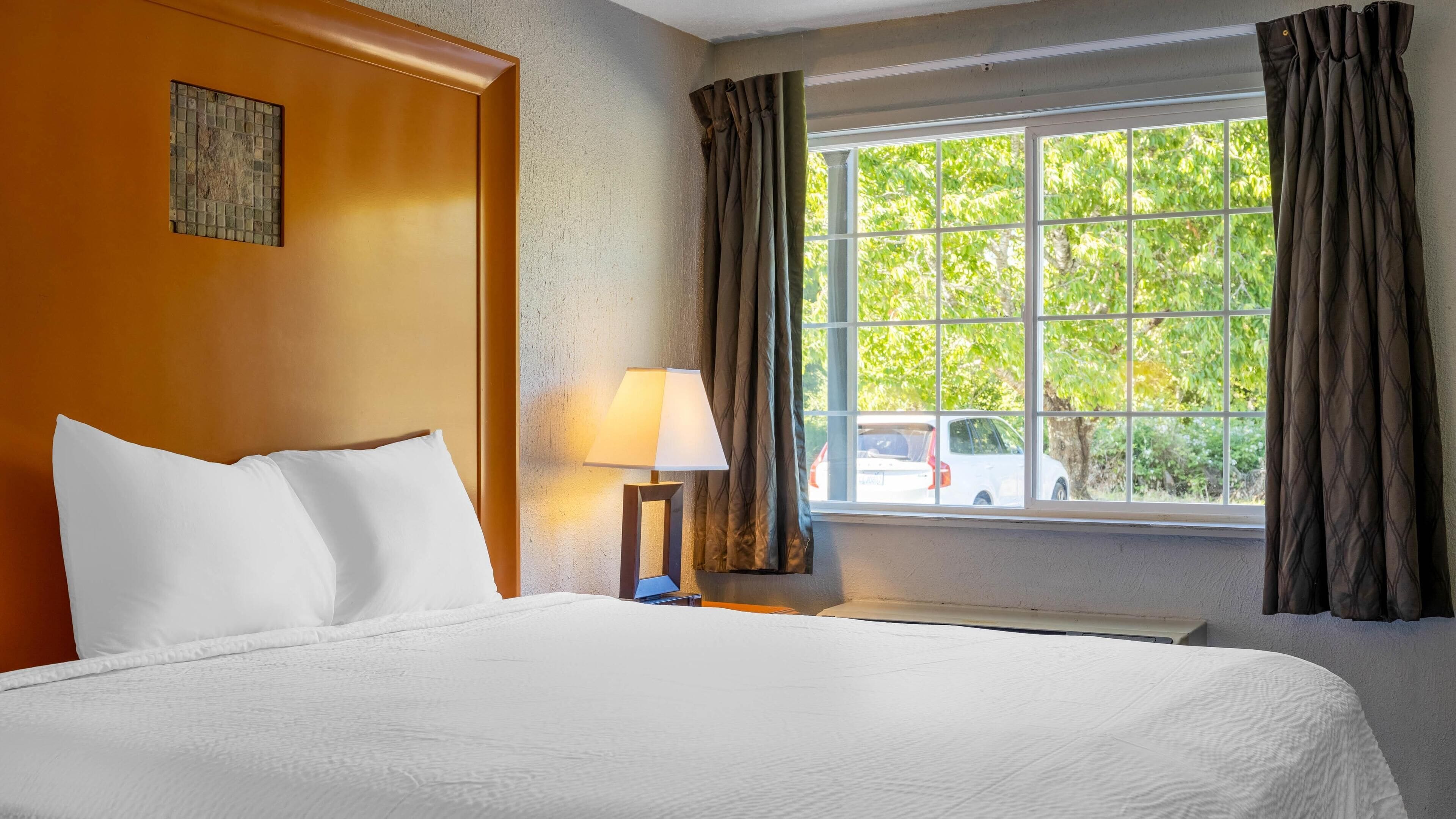 Quarto de hotel com cama grande, janela com cortinas, luminária e vista para o exterior.