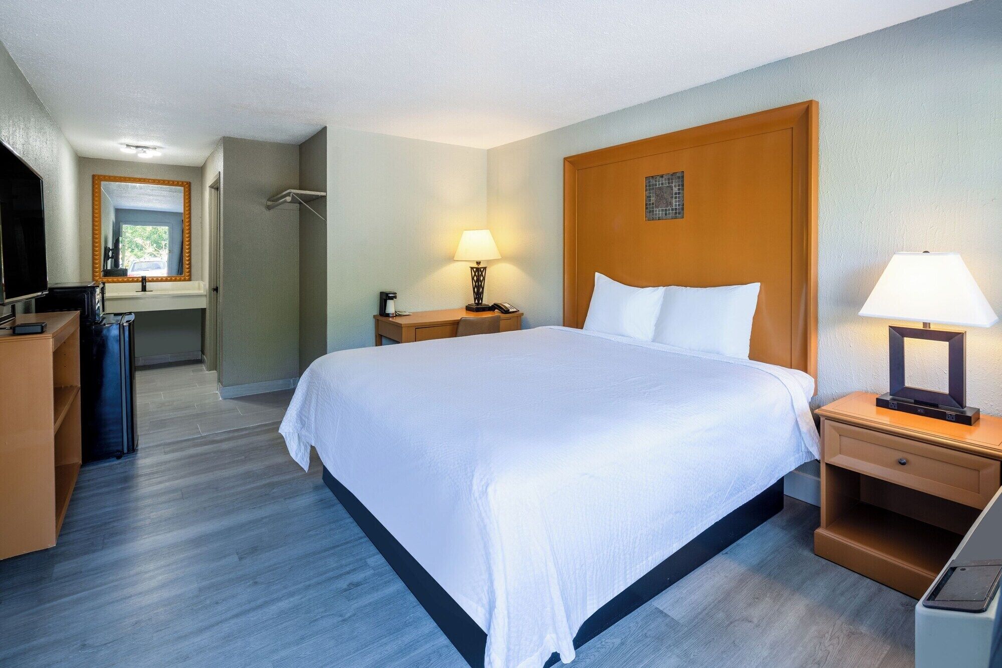 Quarto de hotel com uma cama grande, mesas de cabeceira, escrivaninha, cadeira, espelho e televisão.