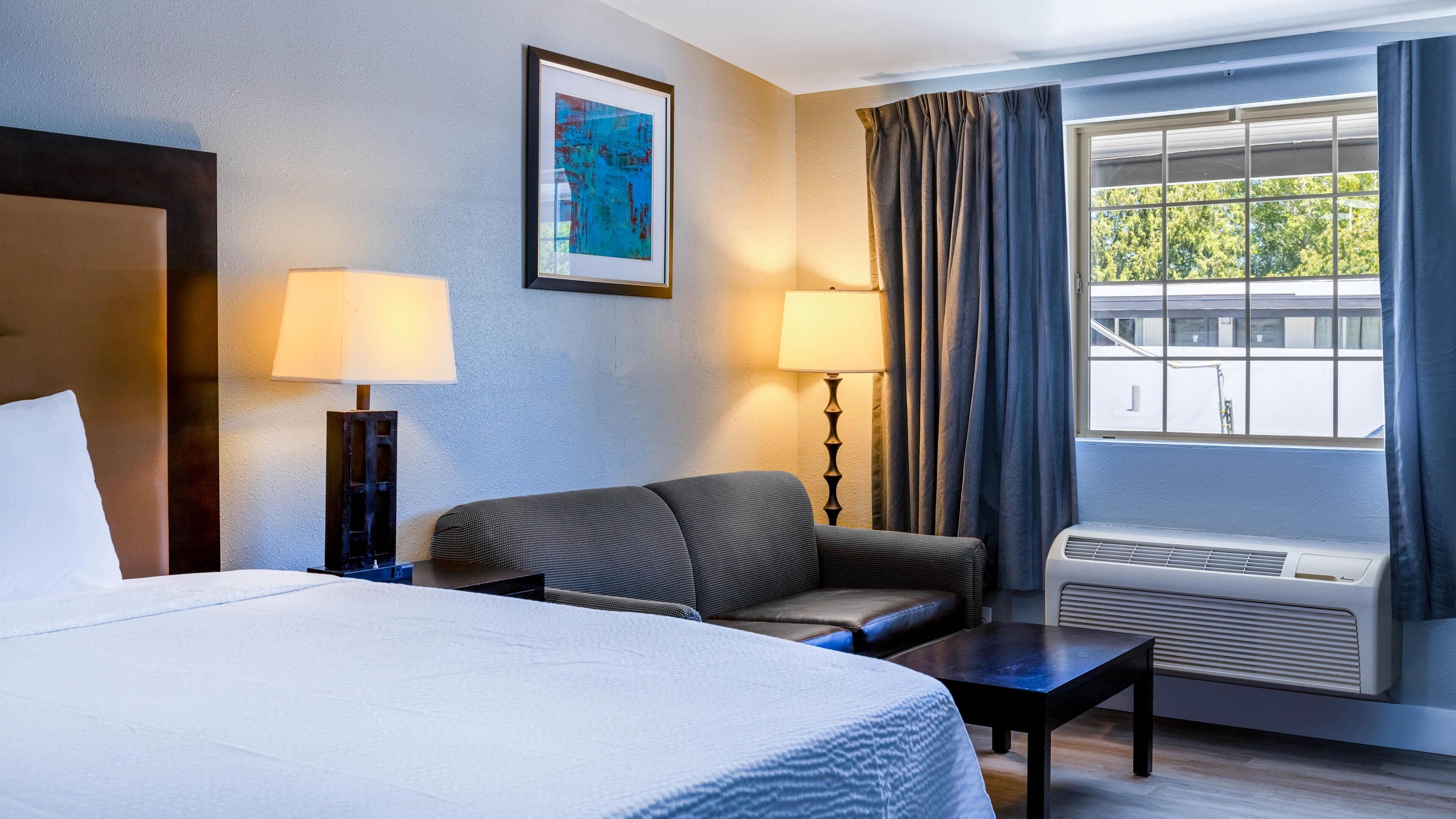 Ein Hotelzimmer mit einem Bett, einem Schlafsofa, einem kleinen Tisch, zwei Lampen, einem Fenster mit Vorhängen und einem Bild an der Wand.