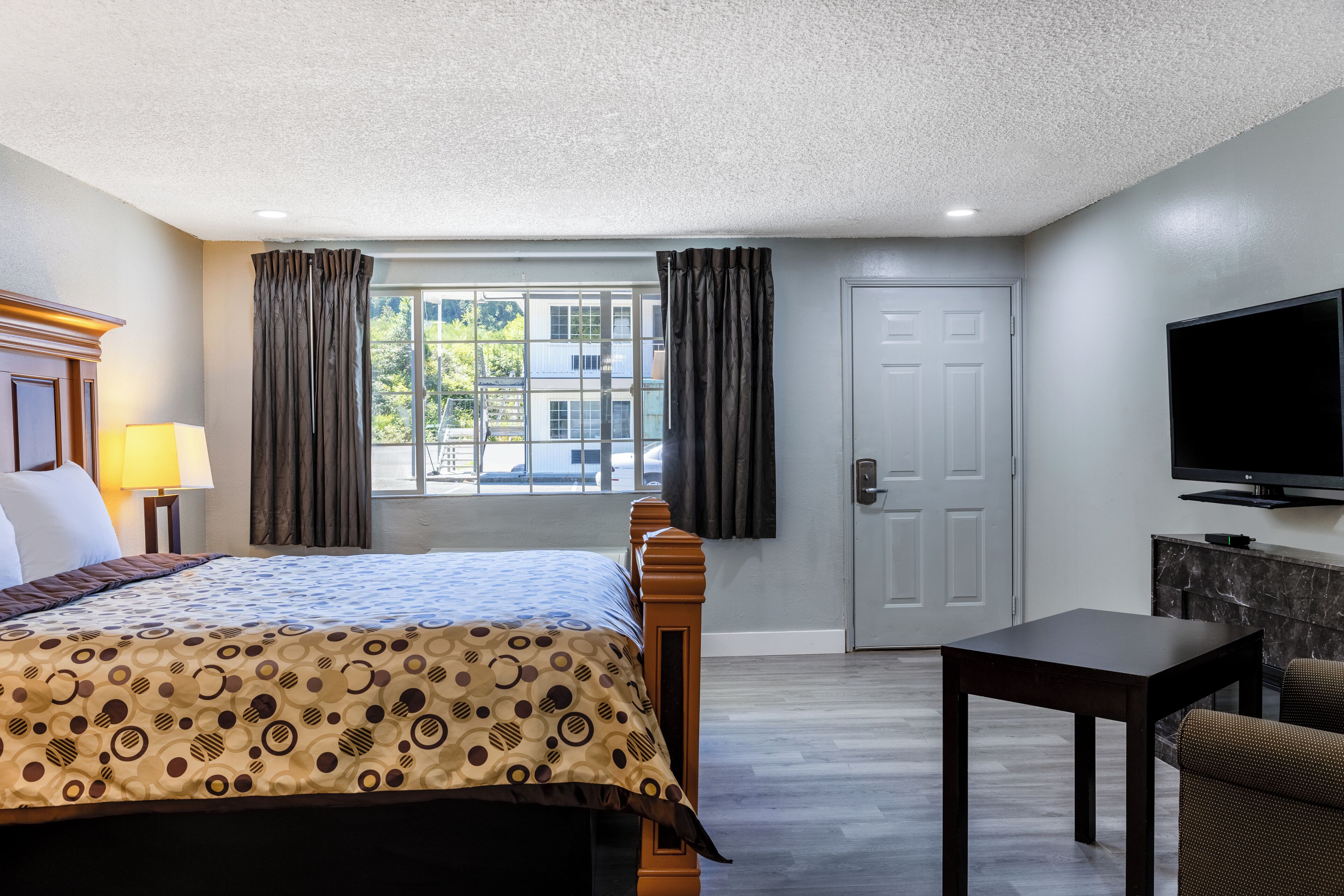 A hotel room with a bed, a TV, a table, and a view of the outside.