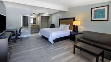 A hotel room with a bed, a desk, a chair, a sofa, a TV, a bathroom, and a painting on the wall.