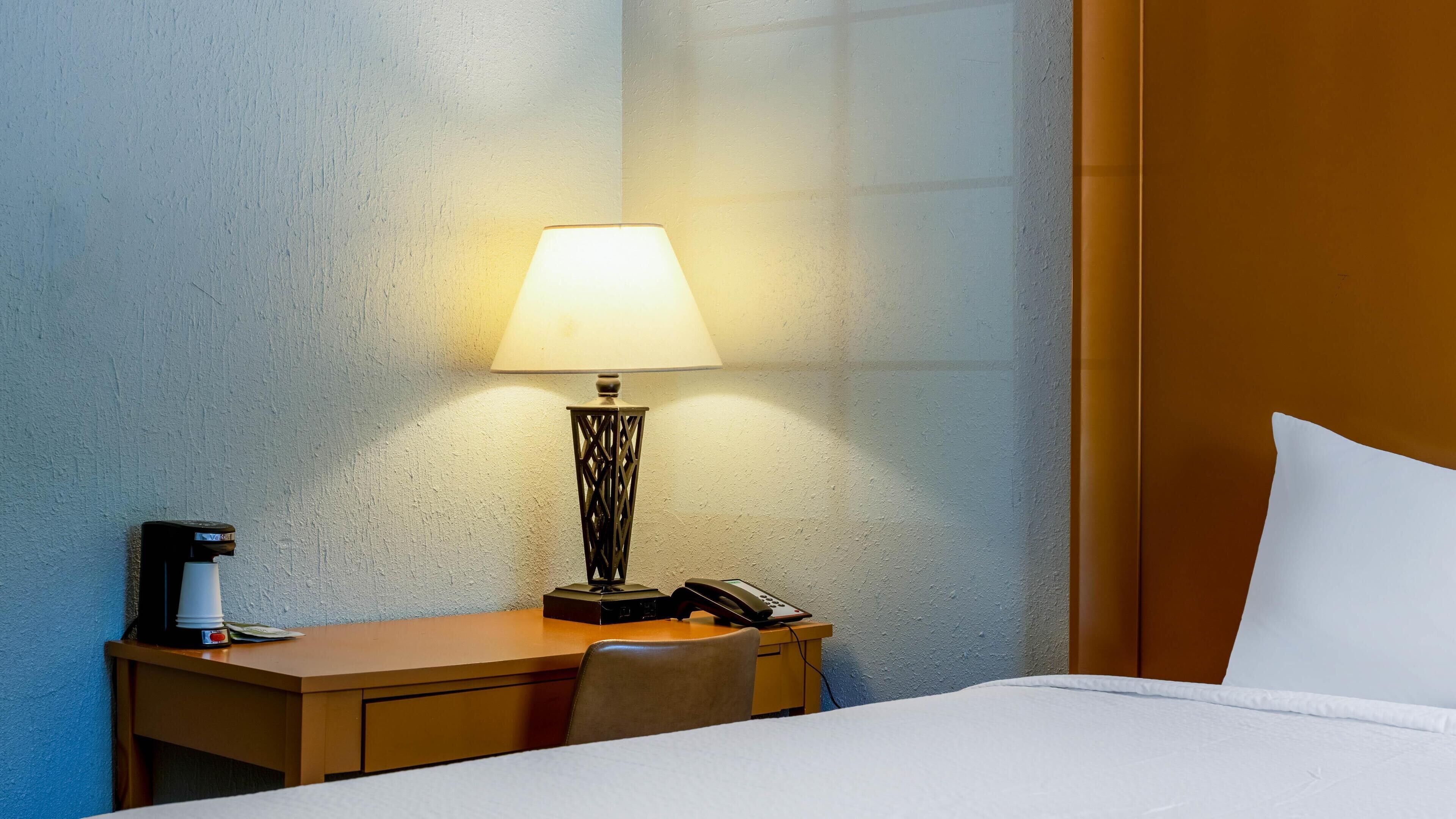 Ett hotellrum med en säng, ett skrivbord med en lampa, en telefon och en kaffebryggare.