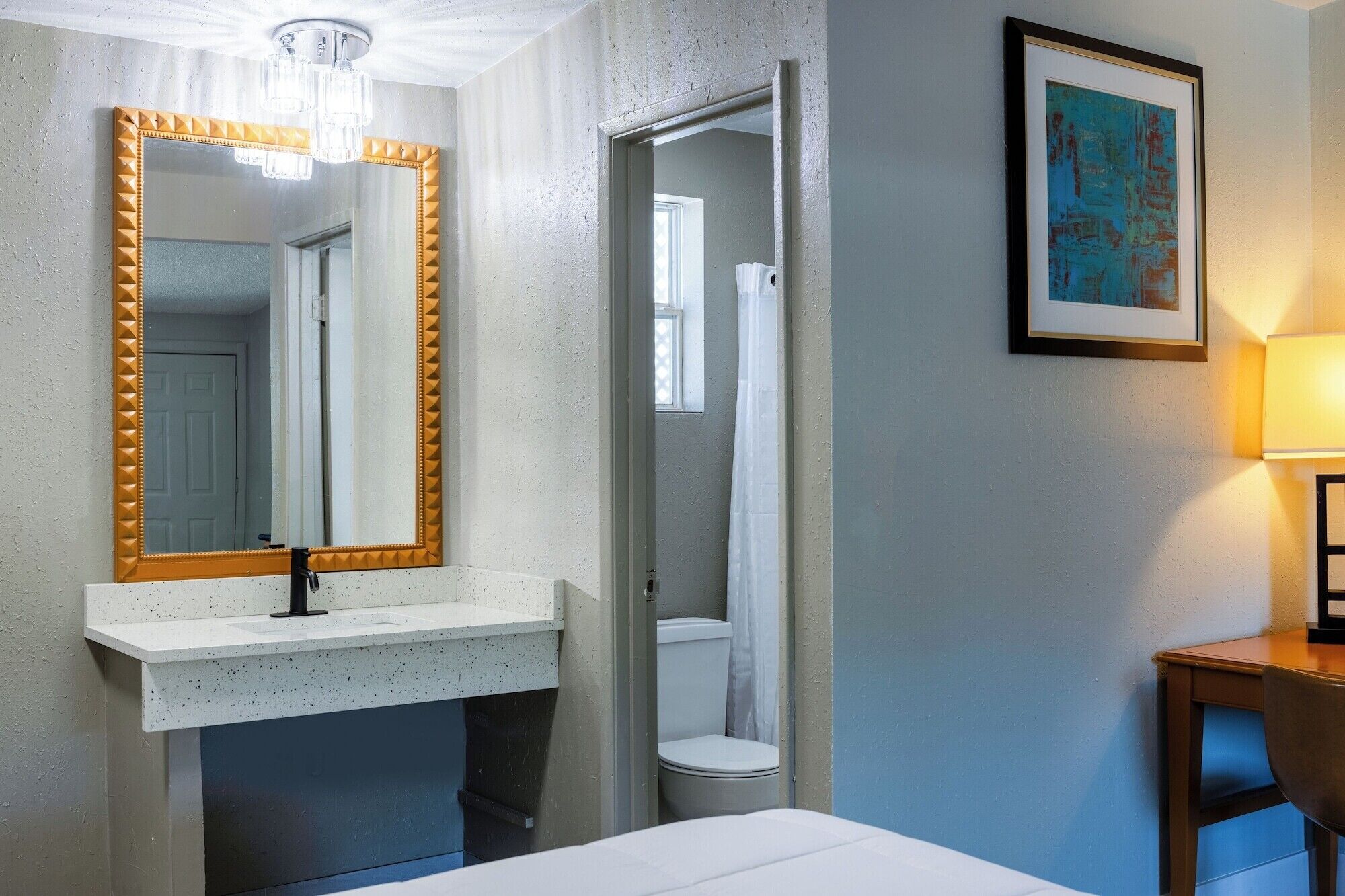 Une salle de bain avec un lavabo blanc, un miroir avec un cadre doré, des toilettes et une douche. Un tableau encadré est accroché au mur et une lampe se trouve sur le côté droit.