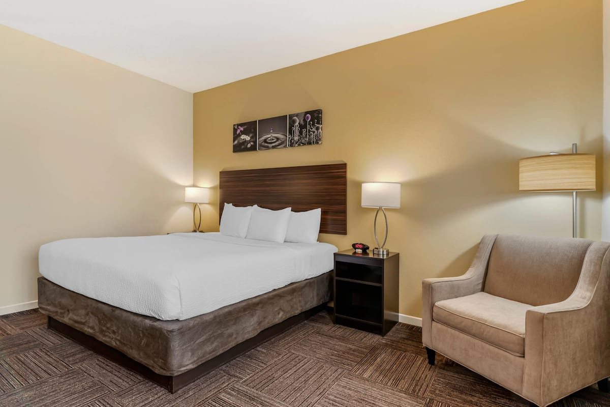 A hotel room with a large bed, two bedside lamps, a nightstand, a beige armchair, and a wall-mounted artwork.