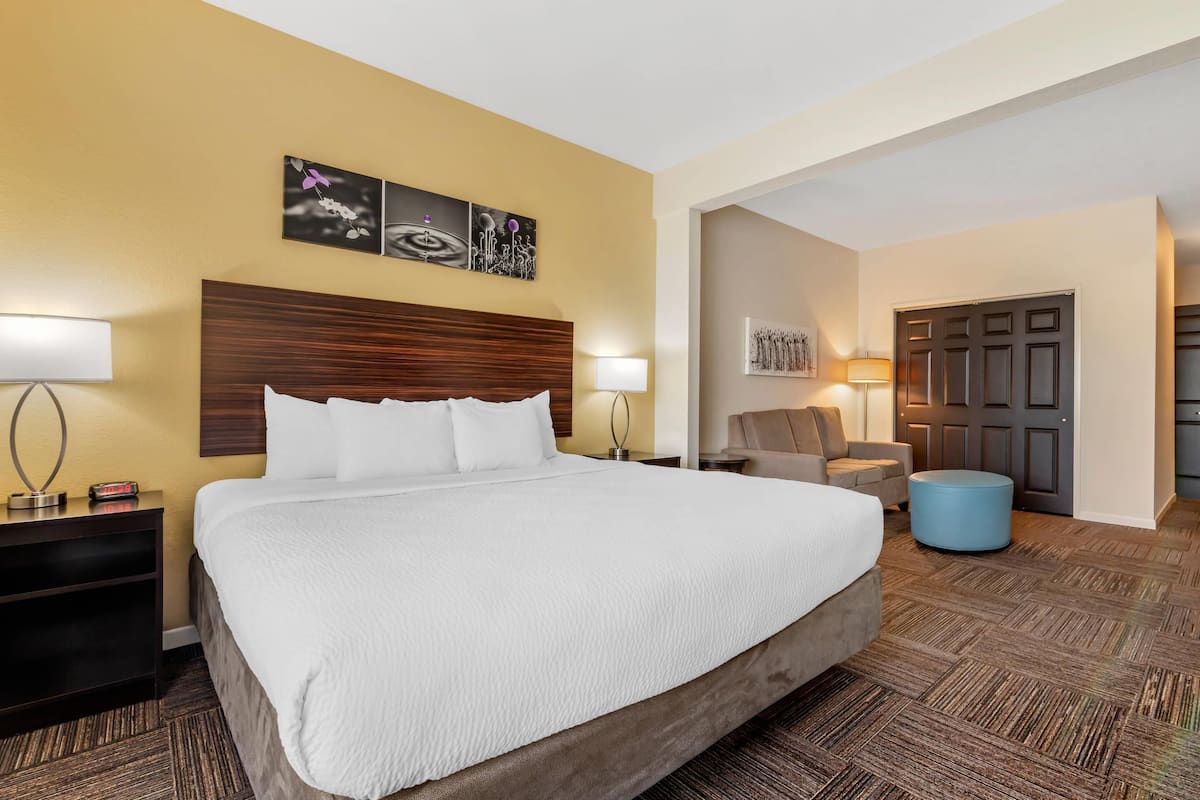 A hotel room with a large bed, a wooden headboard, two bedside lamps, a chair, a blue ottoman, and a door.