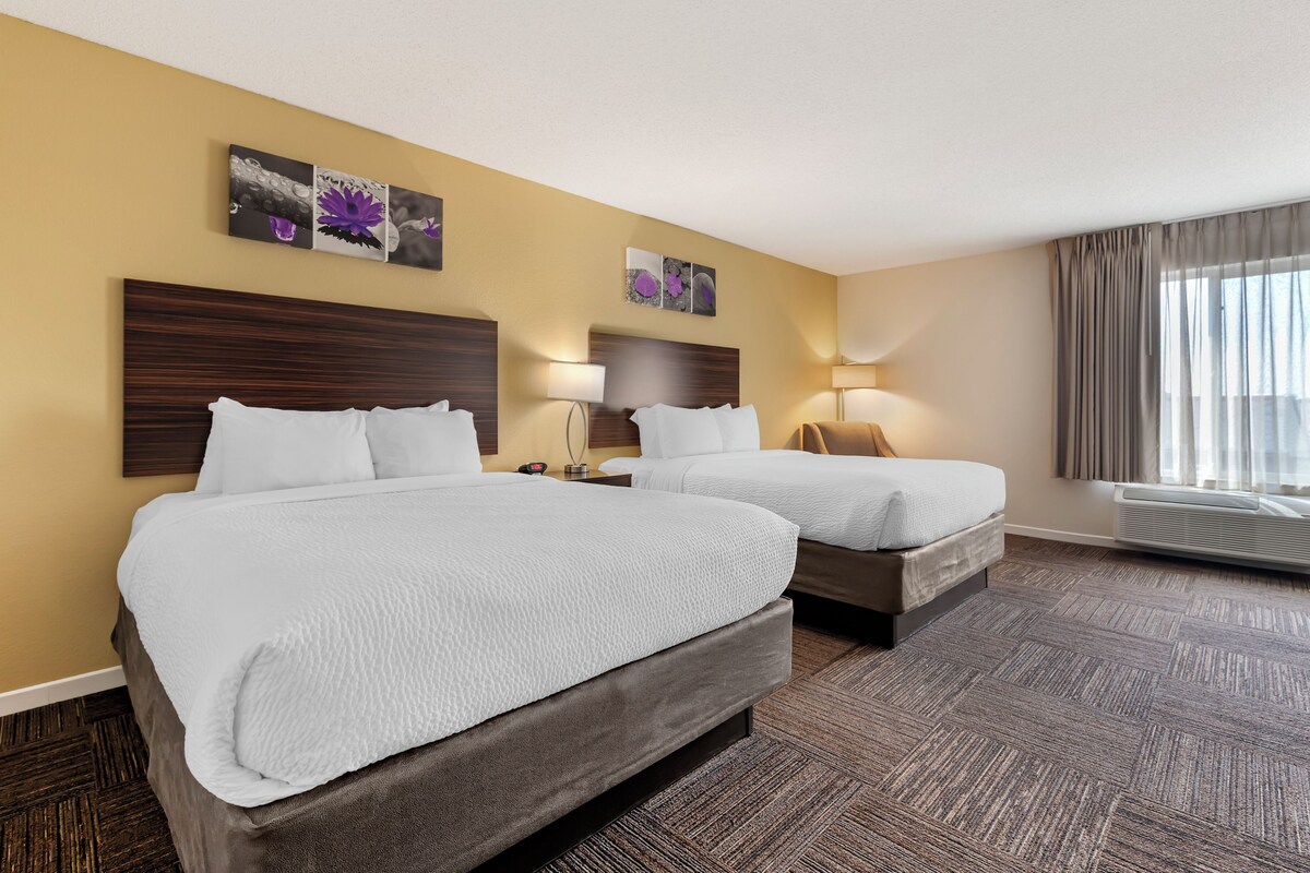 A hotel room with two beds, a wooden headboard, a large window with curtains, and a wall-mounted air conditioning unit.