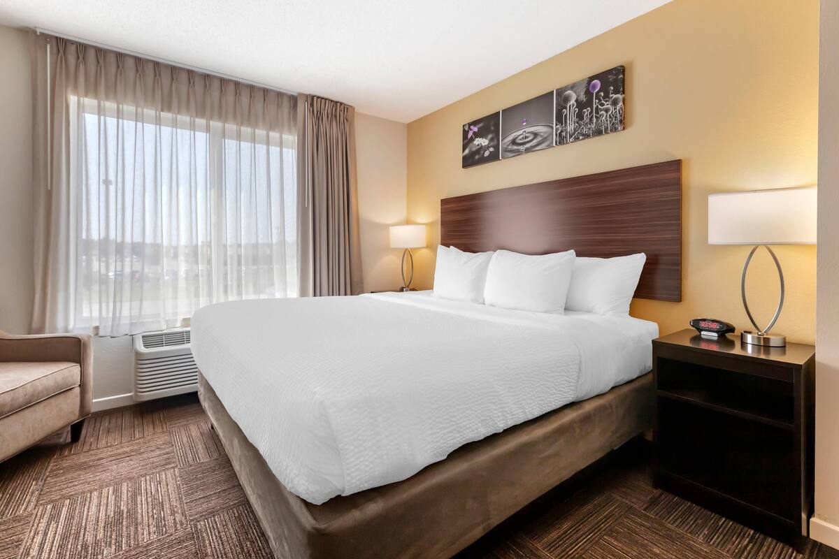 A hotel room with a large bed, a bedside table with a lamp, a chair, and a window with curtains.