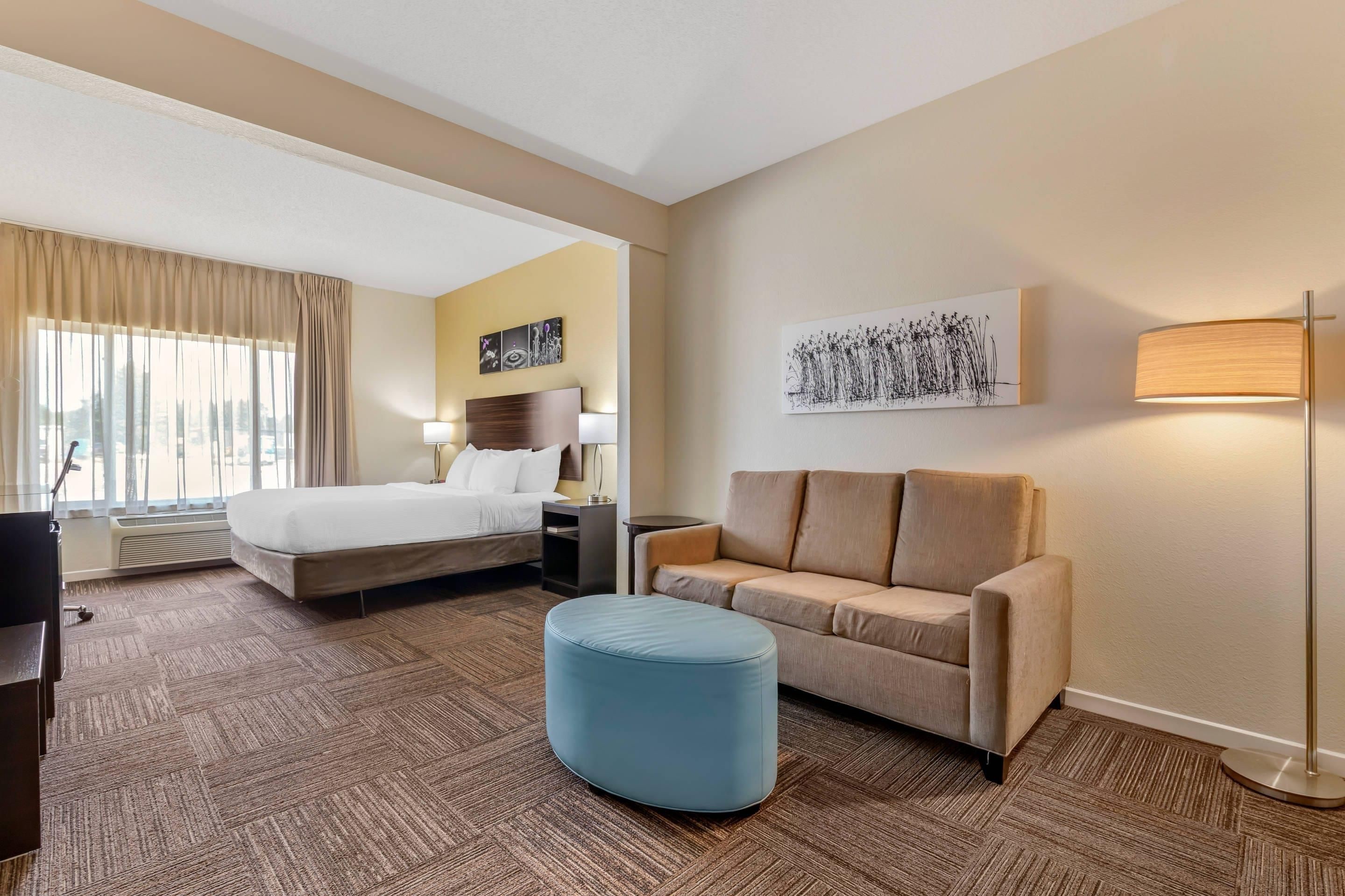 Una habitación de hotel con cama, sofá, reposapiés azul, lámpara y obras de arte en las paredes.