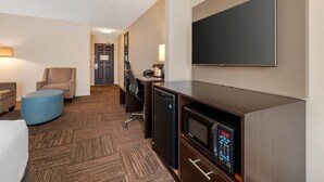 Quarto de hotel com televisão de tela plana, mini-geladeira, micro-ondas, escrivaninha, cadeira e um banquinho azul.