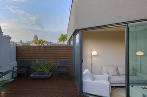 A modern outdoor patio with wooden flooring, a seating area with chairs and a planter, and a glass door leading to a white interior space with a sofa and a lamp.