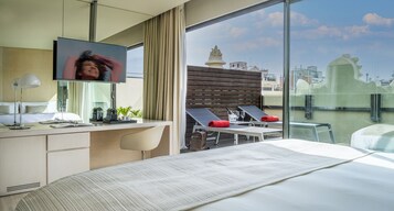 Quarto de hotel moderno com uma cama grande, uma escrivaninha com computador, televisão e varanda com vista para a cidade.