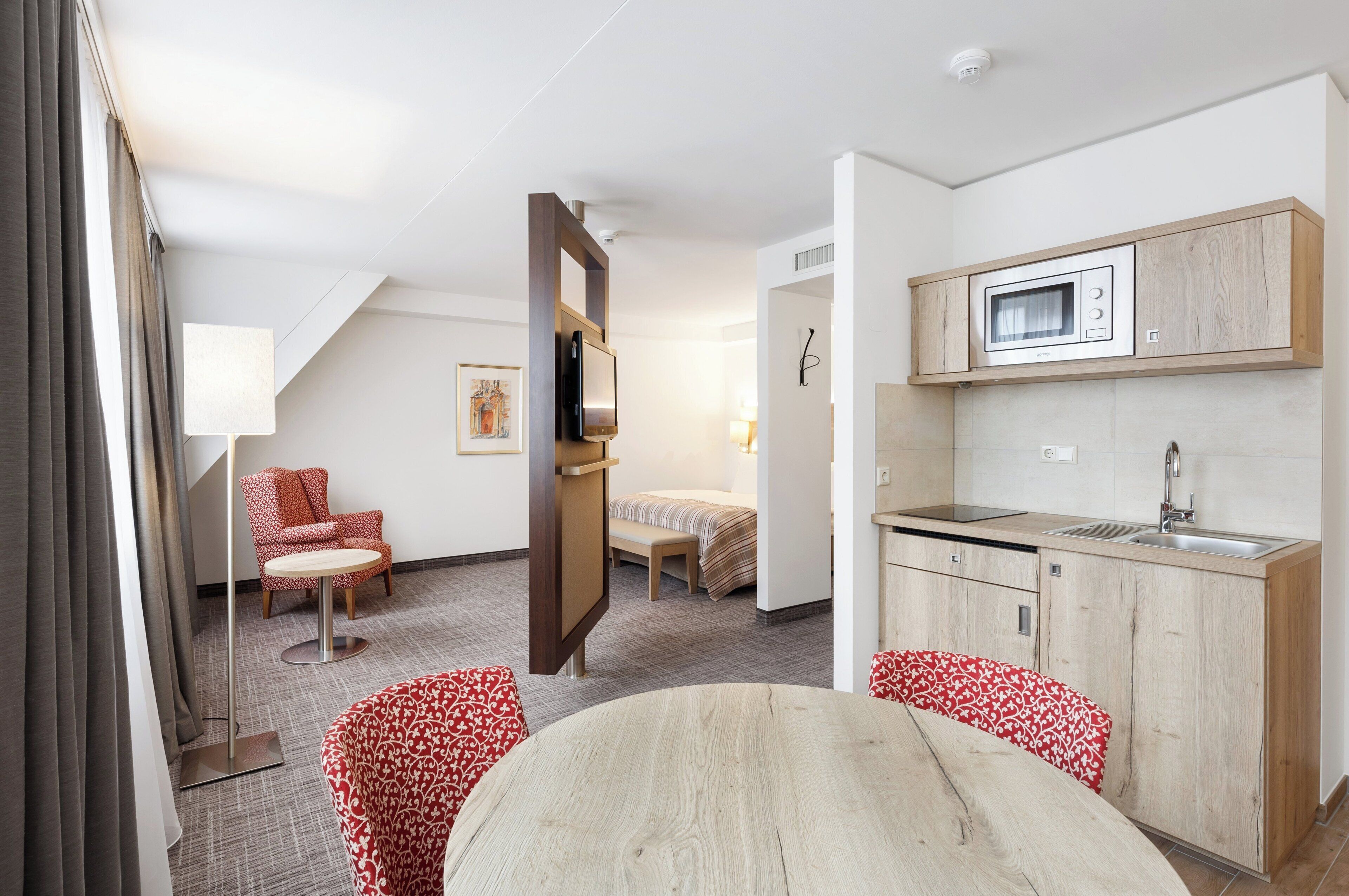 A compact hotel room with a kitchenette, a bed, a seating area with a table, and a TV mounted on a wooden panel.