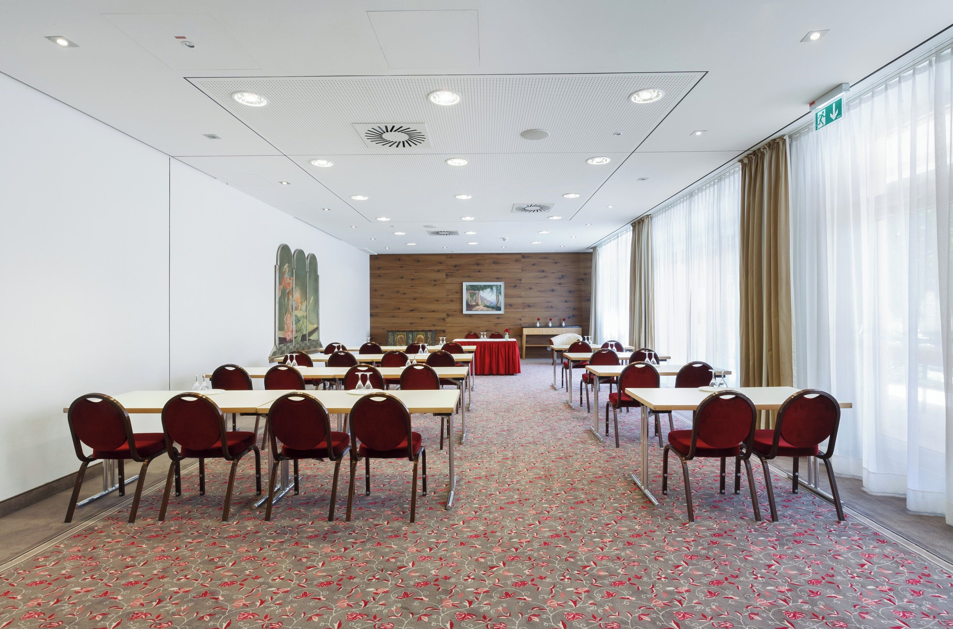 Un salón de conferencias con varias mesas y sillas, una alfombra estampada y un amplio ventanal con cortinas.