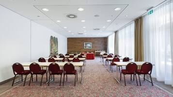 Un salón de conferencias con varias mesas y sillas, una alfombra estampada y un amplio ventanal con cortinas.