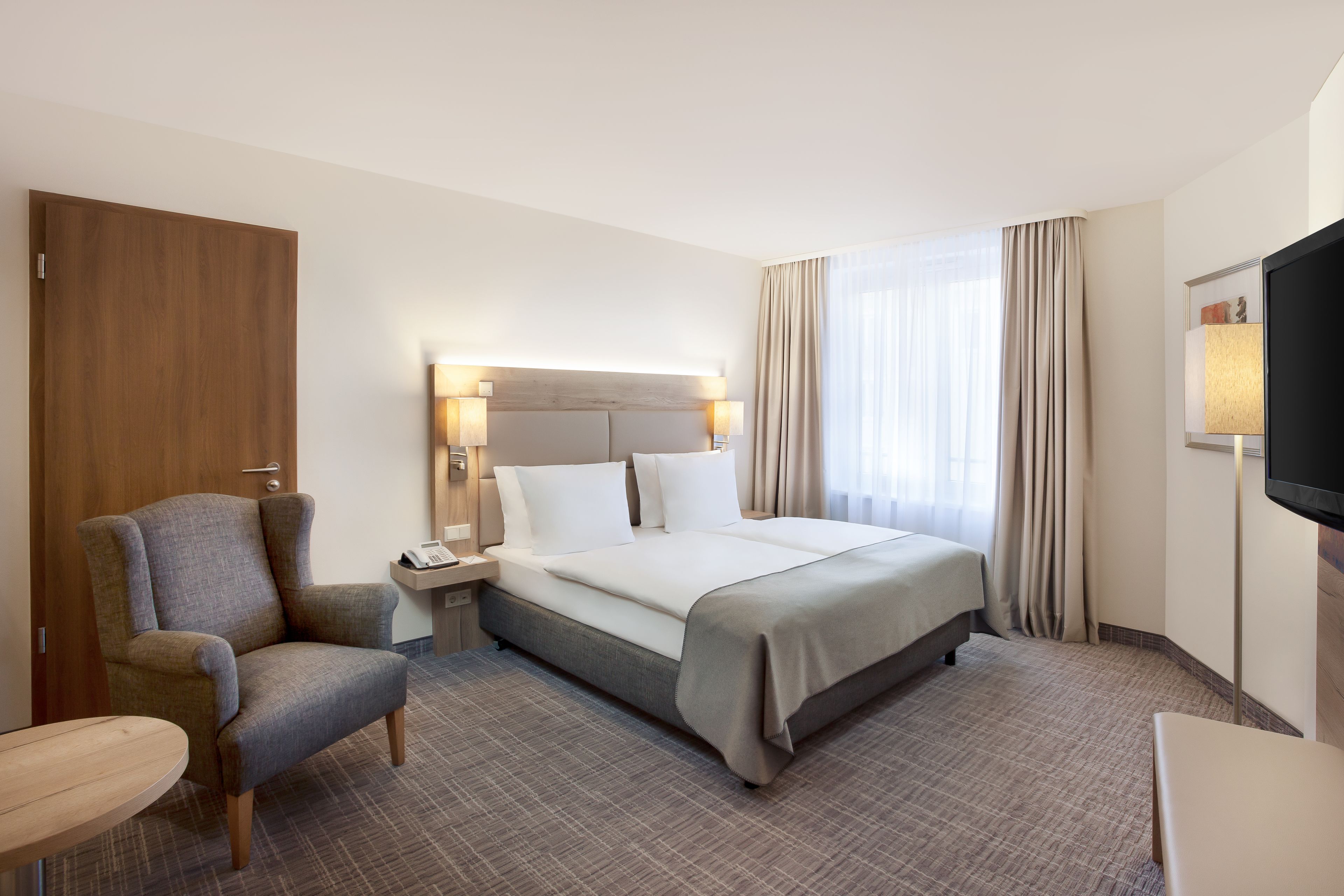 Une chambre d’hôtel avec un lit, une chaise, un bureau, une télévision et une fenêtre avec des rideaux.